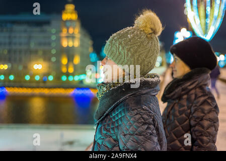 Zwei Mädchen im Profil in warme Winterkleidung stand in der Nacht auf der Straße. Im Hintergrund ist eine festliche Weihnachtsbeleuchtung. Schneeflocken liegen auf dem Stockfoto