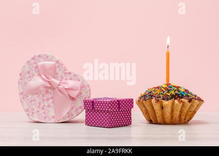 Geschenkverpackung in Form eines Herzens und kleine, leckere Geburtstag Muffin mit Schokoladenüberzug und Karamell, dekoriert mit brennenden festliche Kerze auf Holz- ein Stockfoto