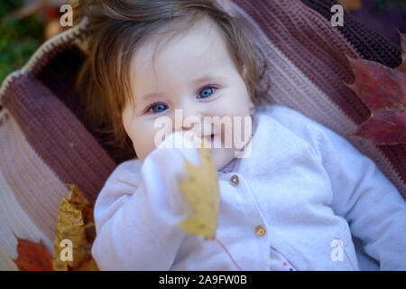 Kaukasisches Baby Mädchen mit blauen Augen und langen braunen Haaren lächelnd und auf dem Rücken auf einer weichen Baumwolldecke in einem herbstlichen Hintergrund Stockfoto