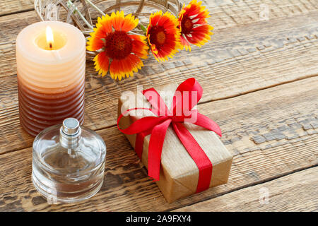 Parfüm, Geschenk oder präsent, in Kraftpapier mit roter Schleife verpackt, brennende Kerze und gelbe Blumen auf Holzplatten. Ansicht von oben. Urlaub Konzept. Stockfoto