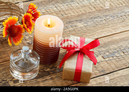 Parfüm, Geschenkbox in Kraftpapier mit roter Schleife verpackt, brennende Kerze und Blumen auf Holzplatten. Ansicht von oben. Urlaub Konzept. Stockfoto