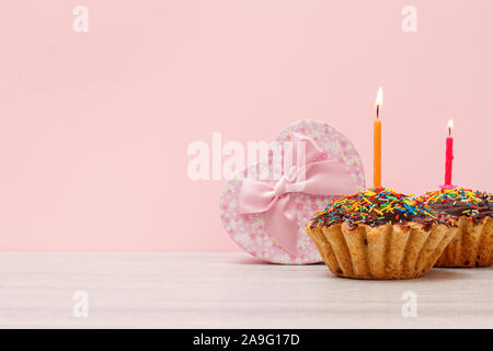 Geschenkverpackung in Form eines Herzens und lecker Geburtstag Muffins mit Schokoladenüberzug und Karamell, mit brennenden festliche Kerze auf Holz- und Rosa bac eingerichtet Stockfoto