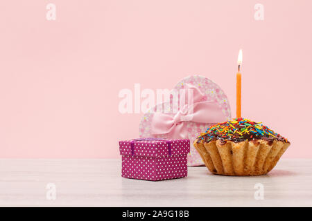 Geschenkverpackung in Form eines Herzens und kleine, leckere Geburtstag Muffin mit Schokoladenüberzug und Karamell, dekoriert mit brennenden festliche Kerze auf Holz- ein Stockfoto