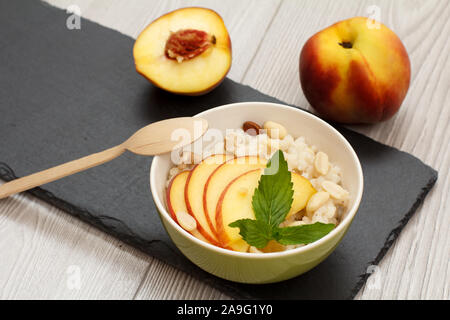 Porzellan Schüssel mit sorghum Porridge, Cashewnüsse, Mandeln, ganz, frisch peache und eine Hälfte, Löffel aus Holz auf Stein. Vegane glutenfreie Sorghum s Stockfoto