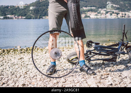 Fahrrad reparieren. Mann Instandsetzung Mountainbike. Radfahrer Mann in Schwierigkeiten hinten Wheel bei Unfall. Mann Bike in der Nähe von See in Italien Hintergrund Stockfoto