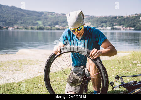 Fahrrad reparieren. Mann Instandsetzung Mountainbike. Radfahrer Mann in Schwierigkeiten hinten Wheel bei Unfall. Mann Bike in der Nähe von See in Italien Hintergrund Stockfoto