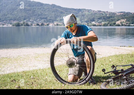 Fahrrad reparieren. Mann Instandsetzung Mountainbike. Radfahrer Mann in Schwierigkeiten hinten Wheel bei Unfall. Mann Bike in der Nähe von See in Italien Hintergrund Stockfoto