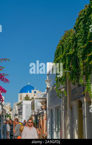 OIA, Santorini, Griechenland - 7. AUGUST 2019; Oia street scene Santorin Blue Dome am Ende und den Tip mit Bougainvillea und Touristen auf der Straße unten. Stockfoto