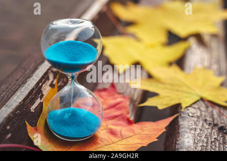 Im Herbst Park auf einer Bank mit Blätter ist Sanduhr mit Blu sand. Herbst Komposition. Die Vergänglichkeit der Zeit Konzept. Bild horizontal Stockfoto