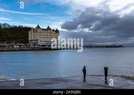 Zwei Jungen Paddeln im Meer und ihre Schuhe vor dem Grand Hotel in Llandudno und Pier bei Sonnenuntergang Stockfoto