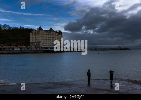 Zwei Jungen Paddeln im Meer und ihre Schuhe vor dem Grand Hotel in Llandudno und Pier bei Sonnenuntergang Stockfoto