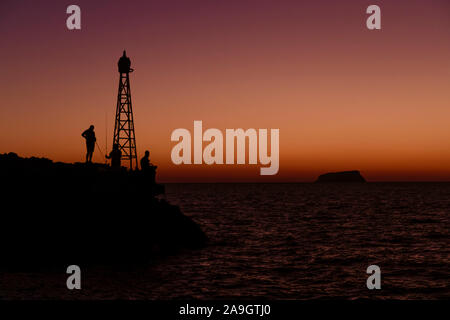 Fischer steht auf Felsen in der Nähe einer Rundumleuchte vor einem leuchtenden Sonnenuntergang auf Santorini in Griechenland. Stockfoto