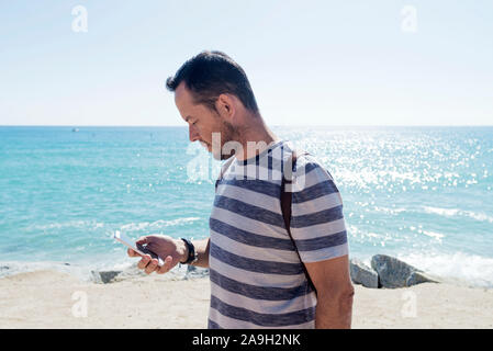 Seitenansicht eines jungen Mann im t-shirt bei der Verwendung von Mobile Phone am Meer in sonniger Tag Stockfoto