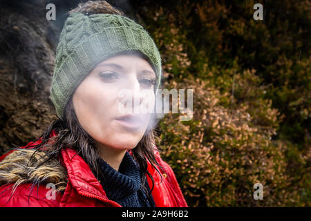 Eine junge Frauen tragen eine warme Mütze und roten Mantel nimmt einen Bruch von Wandern, Bergsteigen eine rauchen, Zigarette Pause zu haben und Ausruhen Stockfoto