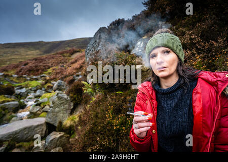 Eine junge Frauen tragen eine warme Mütze und roten Mantel nimmt einen Bruch von Wandern, Bergsteigen eine rauchen, Zigarette Pause zu haben und Ausruhen Stockfoto
