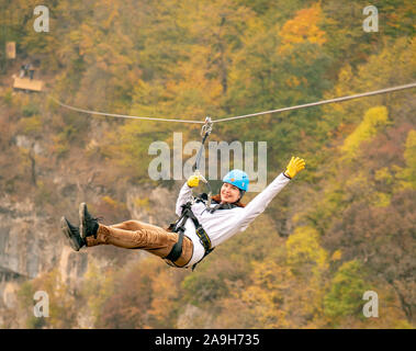 Eine Frau in einem Helm fliegt auf einer Seilrutsche über einen bunten Herbst Schlucht. Stockfoto