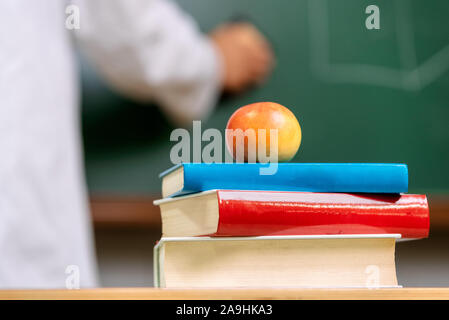 Detailansicht, eines Lehrers Schreibtisch mit einem Stapel Bücher und einen roten Apfel vor einem schwarzen Brett. Stockfoto