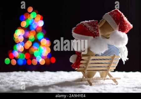 Brauner und weisser Teddybär mit Weihnachtsmütze sitzen auf Holzbank, wenn im Winter schneit auf bunten bokeh Lichter des Weihnachtsbaums. Stockfoto