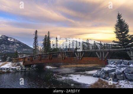 Holzfußgängerbrücke Snowy Spring Creek Dramatischer Sonnenuntergang. Entfernte schneebedeckte kanadische Rocky Mountains Landschaft, Stadt Canmore, Alberta Stockfoto