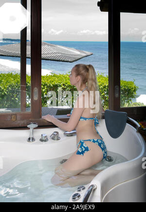 Junge schöne Frau im Badeanzug sitzt in einer großen, modernen Badewanne mit Wasser gefüllt und schaut aus dem Fenster in die Büsche und das Meer Stockfoto