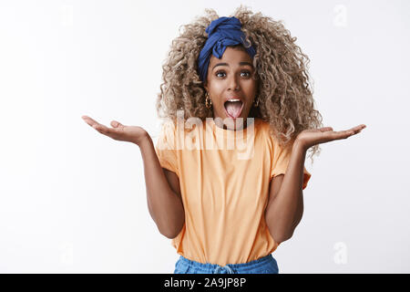 Charismatische gut aussehende african-american blonde lockige Frau in stilvollen orange T-Shirt, Stirnband, amüsiert und schrien: Hände heben seitwärts beeindruckt und ein Stockfoto