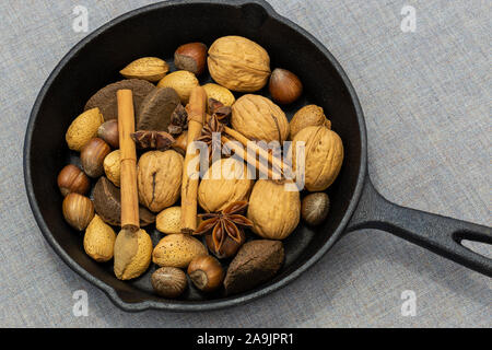 Walnüsse, Haselnüsse, Paranüsse und Walnüsse in einer gusseisernen Pfanne mit Zimtstangen und Sternanis. Auf einem grauen Tischdecke Stockfoto