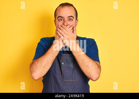 Junge Ladenbesitzer Mann mit Schürze stehend über isolierte gelben Hintergrund schockiert über den Mund mit Händen für Fehler. Geheime Konzept. Stockfoto