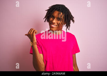 Afro-amerikanische Mann mit Dreadlocks tragende t-shirt stehend über isolierte rosa Hintergrund lächelnd mit glücklichen Gesicht und mit Verweis auf die Seite Stockfoto