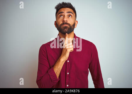 Junge Inder tragen Rot Elegantes Shirt stehen über isolierte grauer Hintergrund Denken konzentriert über Zweifel mit dem Finger auf das Kinn und sah Stockfoto