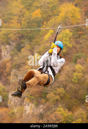 Eine Frau in einem Helm fliegt auf einer Seilrutsche über einen bunten Herbst Schlucht. Stockfoto