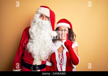 Mittleres Alter Paar Tragen santa Kostüm und Gläser über isolierte gelb hinterlegt angezeigt und zeigt mit dem Finger Nummer eins beim Lächeln konfi Stockfoto
