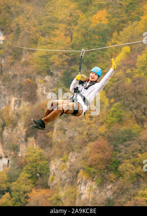 Eine Frau in einem Helm fliegt auf einer Seilrutsche über einen bunten Herbst Schlucht. Stockfoto