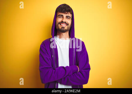 Junger Mann mit Tattoo tragen lila Sweatshirt mit Kapuze über isolierte gelben Hintergrund glücklich lächelnd mit verschränkten Armen in die Kamera schaut. Stockfoto