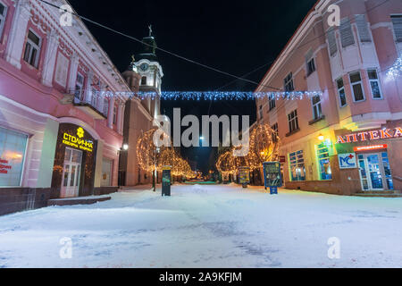 Uschhorod, Ukraine - 06 Jan, 2019: Winter Nacht in der Stadt. Wunderschöne blaue und gelbe Weihnachtsbeleuchtung. Leere Voloshyna Straße im Schnee bedeckt. Cath Stockfoto