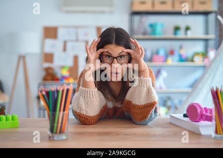 Junge schöne Lehrerin Frau mit Pullover und Gläsern sitzen am Schreibtisch im Kindergarten versuchen, Augen mit den Fingern, schläfrig und müde für morni zu öffnen. Stockfoto