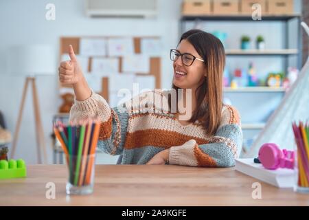 Junge schöne Lehrerin Frau mit Pullover und Gläsern sitzen am Schreibtisch im Kindergarten Suchen stolz lächelnd tun Daumen nach oben Geste zur Seite Stockfoto