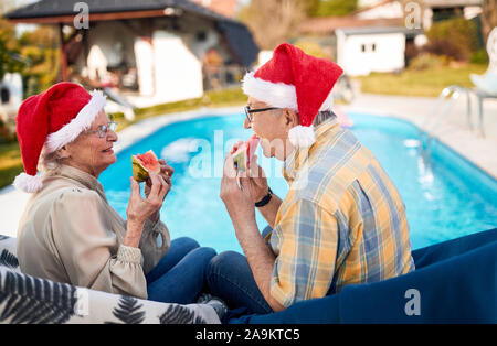 Gerne älterer Mann und Frau in Santa Hüte auf Weihnachten und essen Wassermelone Stockfoto