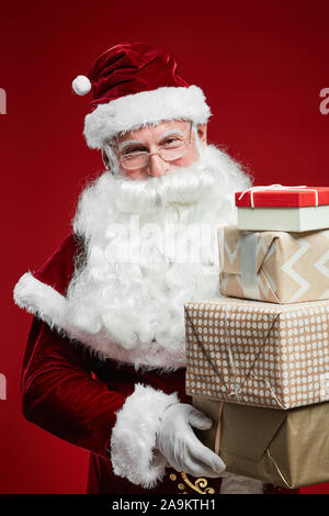 Portrait von Santa Claus rot Kostüm und Hut holding Haufen Weihnachtsgeschenke isoliert auf rotem Hintergrund Stockfoto