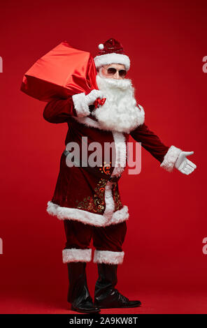Portrait von Santa Claus in Sonnenbrille mit roten Sack mit Geschenken, stehend gegen den roten Hintergrund Stockfoto