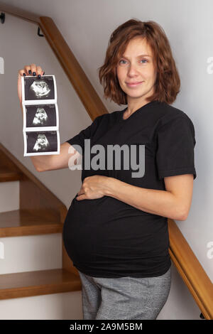 Bild von Schwangeren lächelnd kaukasischen curly Frau sitzt auf der hölzernen Treppen und posiert und zeigt Ultraschalluntersuchungen auf ihrem Bauch auf weißem Hintergrund. L Stockfoto