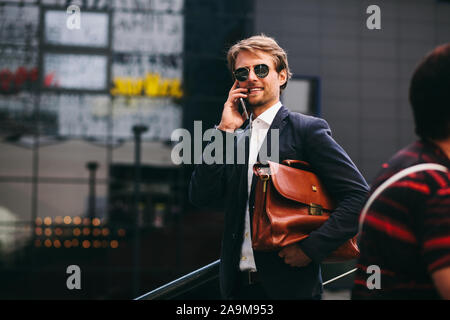 Ein Mann mittleren Alters, der arbeitet in einem Büro ist zu Fuß entlang der Straße mit einer Aktentasche in der Hand und sprechen für eine Mobile und lächelnd Stockfoto