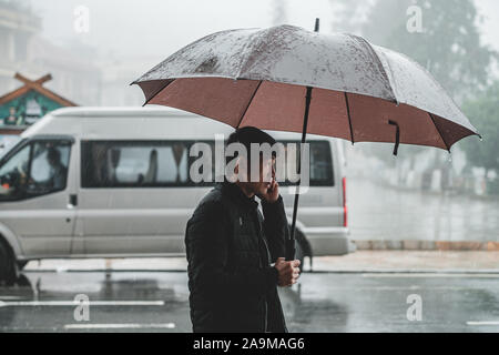 Sapa, Vietnam - 13. Oktober 2019: ein asiatischer Mann mittleren Alters an der Kamera im Regen unter einem Sonnenschirm in der kleinen Bergstadt Sapa Stockfoto