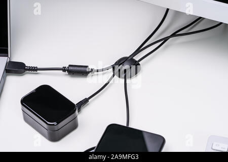 Nahaufnahme der organisierten Kabelhalter am weißen Tisch mit Ladekabel angeschlossen zu drahtlosen Geräten Stockfoto