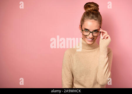 Ein Mädchen mit einem Fernlicht steht in einem Pullover auf einem rosa Hintergrund, lächelt und schaut auf die Seite, halten Sie Gläser mit ihrer Hand Stockfoto