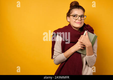 Eine Brünette mit Brille und ein Burgunder Schal, mit einem hohen Bun aufkreuzt, um ein Buch zu sich selbst und schaut verträumt auf. Stockfoto