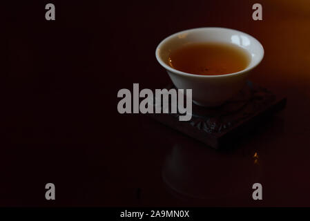 Eine kleine weiße Chinesische Teetasse mit schwarzen frischen Kaffee in Es steht gegen Braun und dunklen Hintergrund und ist in goldenes Licht auf einer Tischplatte mit s wider Stockfoto