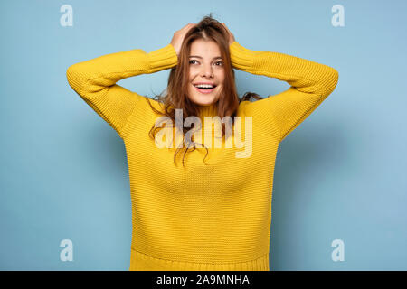 Die brünette steht auf einem blauen Hintergrund in einem gelben Pullover und schaut auf die Seite, ihr Haar ruffling mit ihren Händen. Stockfoto