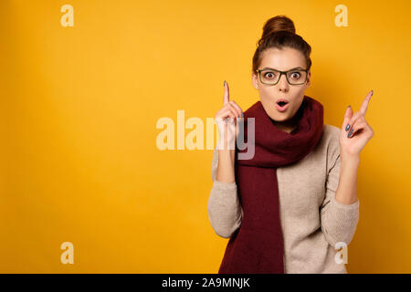 Eine Brünette mit Brille und ein Burgunder Schal, mit einem Fernlicht, steht mit einem überraschten Gesicht und Finger nach oben. Stockfoto