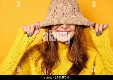 Eine Brünette steht auf einem gelben Hintergrund in einem gelben Pullover, ziehen Sie einen Hut über eine halbe-shirt und Lächeln Stockfoto