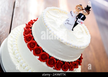 Weisse Hochzeitstorte mit Braut und Bräutigam Figuren an der Spitze. Schöne weisse Hochzeitstorte mit roten Rosen in der Tabelle mit den kleinen Mann und Frau Marionetten Stockfoto